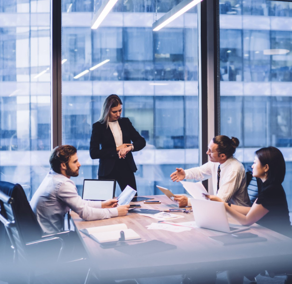 Ambiente Corporativo composto por 4 pessoas sendo 3 sentadas e 1 em pé, vestidas com roupas sociais, sobre a mesa notebooks e objetos