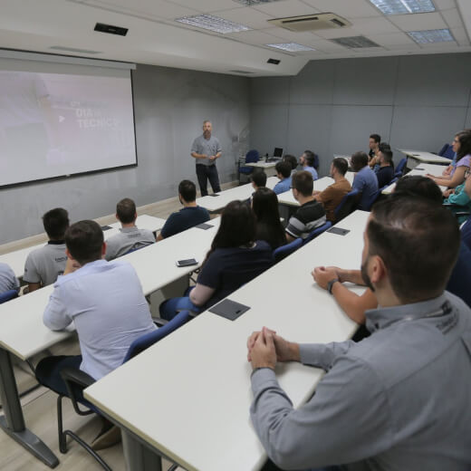 Homem dando treinamento para funcionários que estão sentados atras de algumas mesas brancas e a frente tela branca com projetor