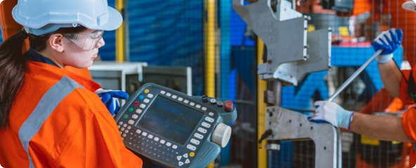 Colaboradores em ambiente industrial manuseando máquinas, com uniforme laranja de faixas cinzas, luvas azuis, capacete branco e óculos de proteção