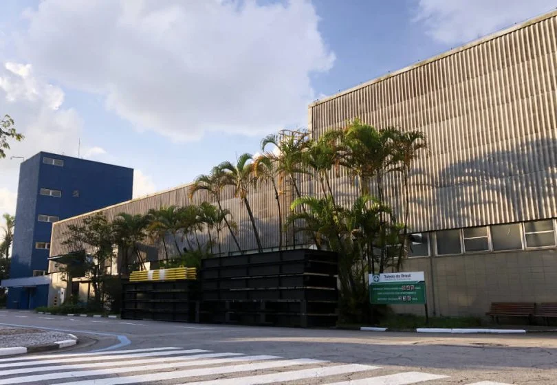 Empresa Toledo do brasil possui estrutura robusta cinza, alguns coqueiros na frente e placa de identificação da empresa na cor verde