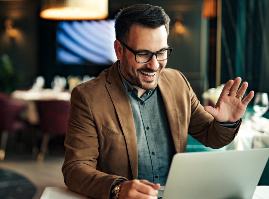 Homem em Reunião, vestido com blazer marrom, camisa cinza, usa óculos de grau, fazendo sinal de cumprimento em frente ao notebook