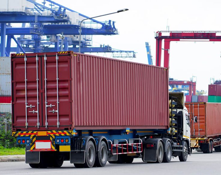 Caminhões com containers alinhados em porto brasileiro, ilustrando desafios logísticos e transporte de cargas.