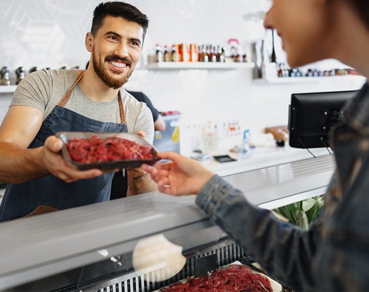 Homem sorridente atrás do balcão de uma loja entrega uma bandeja com carne fresca a uma cliente, simbolizando atendimento próximo e de qualidade.