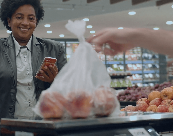 Cliente acompanhando a pesagem de frutas em balança no setor de FLV de um supermercado.